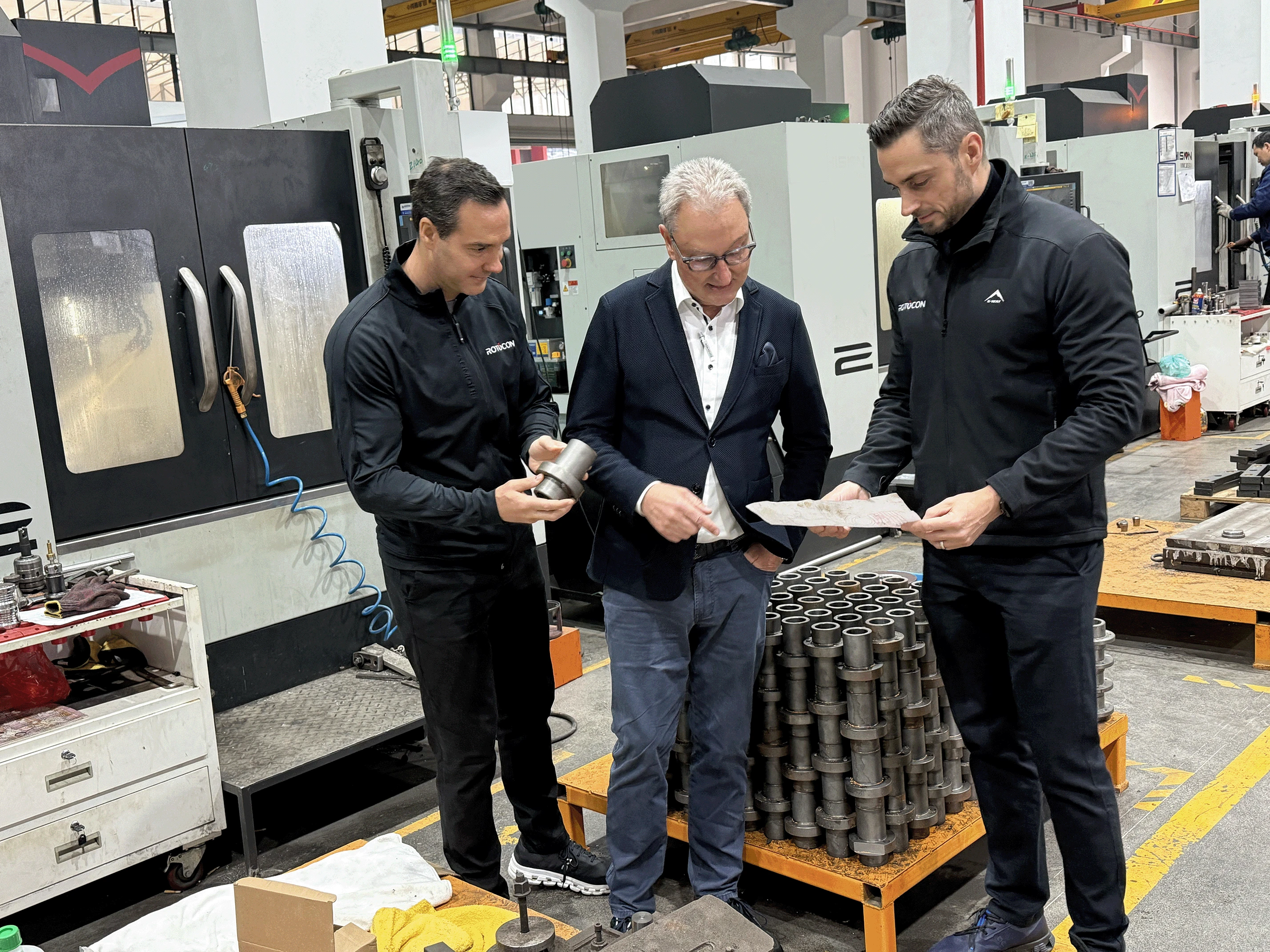 Three men in a manufacturing facility discussing metal parts, one holding a component and another examining a drawing.